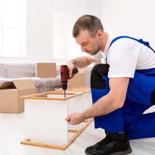 Handyman In Blue Coverall Uniform Using Electric Drill Making Furniture Assembly, Installing Shelf Indoors. Professional Carpenter Working Fixing Cabinet. Carpentry Concept