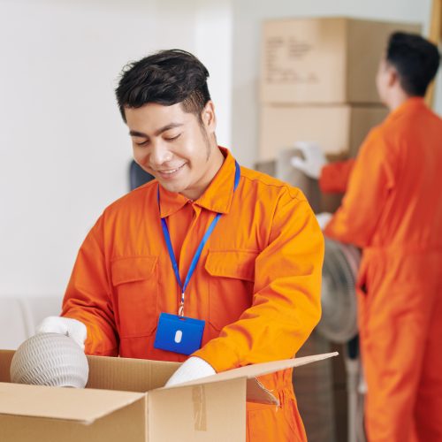 Smiling Vietnamese moving service worker packing fragile objects in cardboard boxes